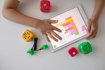 Flat-lay photograph of brightly colored toys on a white tabletop, and a child's hands touching an iPad
